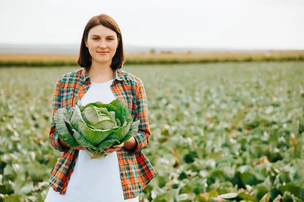 stock image Front view copy space young farmer woman standing field with cabbage in her hands and smiling sweetly. The horizon line is visible in the distance. Blurred background, greenery. Looking at camera.