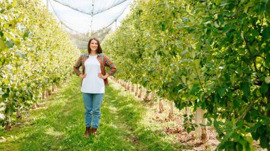 Ön manzara, güzel kadın çiftçi, elma bahçesinde durup güzel kollu kalçalara gülümsüyor. Kadın tarım işçisi günlük kıyafetlerini giyiyor. Kameraya, esmer kadına, küçük işletmelere bakıyor..