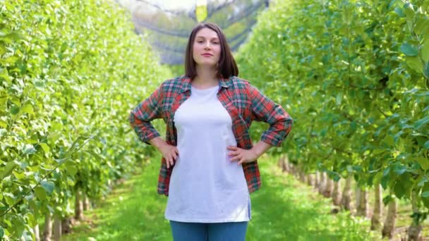フロントビュー若い女性の農家は 腰に彼女の手で果樹園内のリンゴの木の間に立っている 木の列の間にカメラを見ている若い農学者の女の子 — ストック動画
