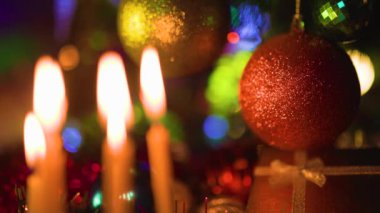 Çerçevede, yanıp sönen ışıkların parlak arka planına karşı Noel toplarının yanındaki mum ateşi. Kırmızı ve sarı parlak Noel topları. Kapat, seçili odak.