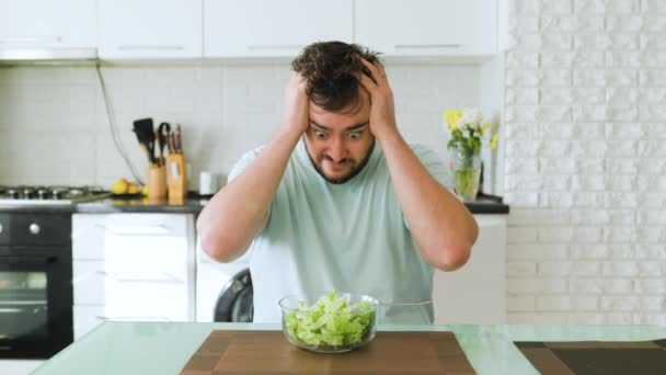 台所に座り 手で頭を抱えていた青年は 必死にカメラを覗き込む レタスと葉物野菜を食べるのに疲れている 健康的な食べ物や好きな料理 健康食品のコンセプト — ストック動画