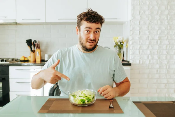 A young dieting man points his index finger at a bowl of salad in front of him, asking Is this supposed to be eaten Green salad healthy food concept. Sits in his kitchen. Looking at camera.