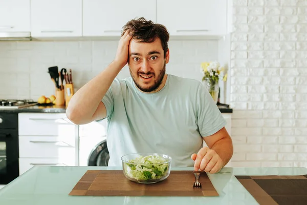 stock image Front view looking at the camera a young guy holds his head in front of him lies a fork and a plate of salad. The action takes place at home in the kitchen. Modern interior and modern food. Stop diet.