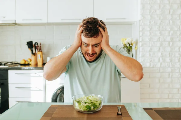 Mladý Muž Sedící Kuchyni Zděšeně Dívá Zelený Salát Který Měl — Stock fotografie