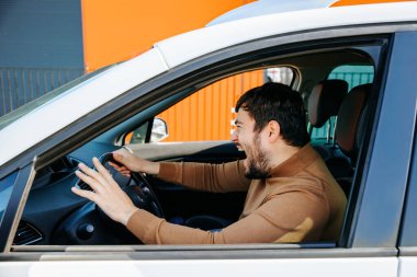 Orta yaşlı beyaz erkek şoför arabanın içinde ve öfkeyle bağırıyor. Erkek, ağzı açık, gergin ve elleriyle el kol hareketi yapıyor. Yan profil