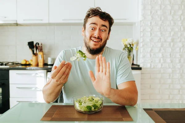 A young guy in a hysterical state renounces vegetables and greens, showing stop with his hand. Front view guy at home in the kitchen. Healthy food. Stop diet.