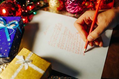 Noel 'de insan eli beyaz kağıda kırmızı kalemle tanınmaz hale gelir. Noel Baba 'ya renkli çelenklerin bulanık arkaplanı etrafında bir mektup yazar. Seçici odaklanma. Kapat..