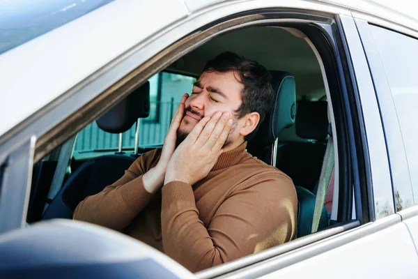 A young driver is holding his cheeks with his hands, his teeth hurt. A lot of time behind the wheel, hard work of the driver. Toothache does not allow to live normally.