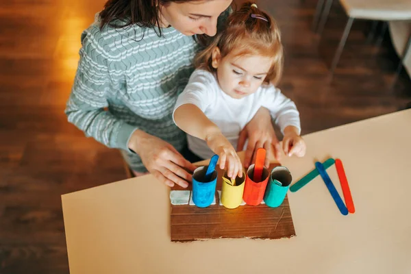 Focused little girl playing a young mother doing experiments with rainbow colors in a modern kitchen together. Happy mother brings up her little daughter at home, developing her cognitive processes.