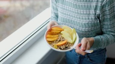 Meyve, tahıl, yulaf ezmesi ve diğer doğal malzemelerle dolu bir tabakta ev yapımı granola tutan bir kadının ellerine yakın çekim. Sağlıklı kahvaltı, karbonhidrat ve kaba liflerle dolu bir kaseye odaklan..