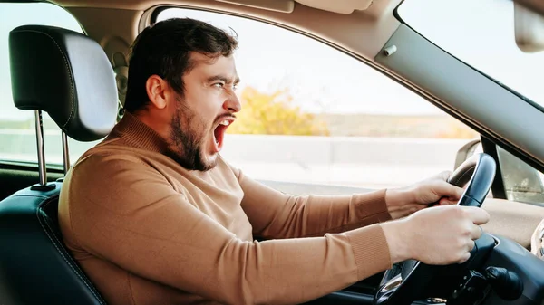 A young male driver screams loudly while sitting at the wheel and looks somewhere on the road. A lot of bad things happen in the life of a driver. There are many troubles on the road.
