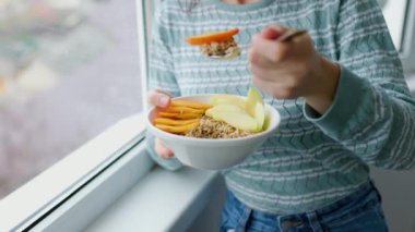 Genç bir kadın müsli kahvaltısı yapıyor. Beyaz bir kasede çiğ meyvelerle birlikte. Doğrudan yukarıdan görülüyor. Kadın bir kaşık dolusu fiber tanecikli taze kaşık tutuyor. Yukarıdan görünüyor. Üst görünüm.