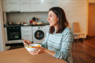 A happy young woman sits at a table, look into the distance, holds a plate with breakfast and a spoon in her hands. Good morning started with healthy food. The girl follows the figure and enjoys life.