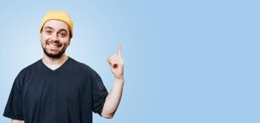 A laughing guy in a yellow cap, in a studio shot with a blue background, with the index finger up he has an idea, eurek. expression joy while also looking at the camera with ample copy space.