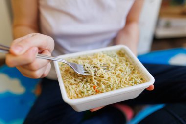 A close-up of hands showing the consumption of junk food showing instant noodles. with a fork in the foreground.