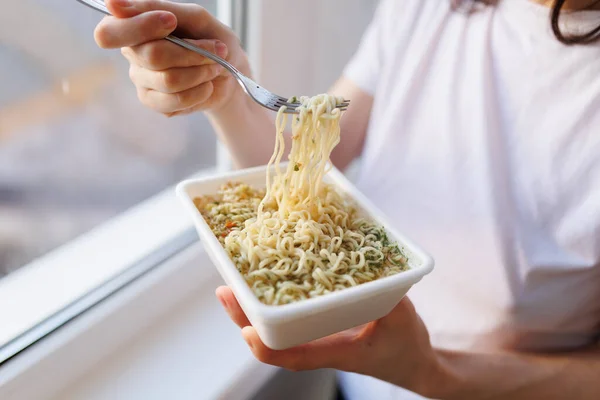 The hands of someone eating a fast food meal of instant noodles, captured in a close-up shot. The fork is in motion, and the scene takes place at home by the window in the kitchen.
