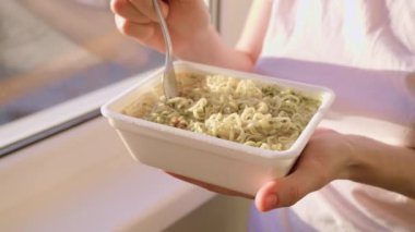 A close-up shot of a hand holding a fork, into a plate of instant noodles, moment of a quick and unhealthy meal eaten at home by the window in the background.
