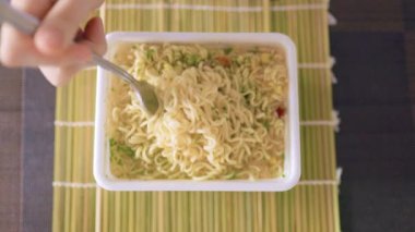 A shot of someone enjoying a quick and unhealthy meal of instant noodles, with the focus on the fork in their hand. with a fork in hand and a plate of fast food in front of them.