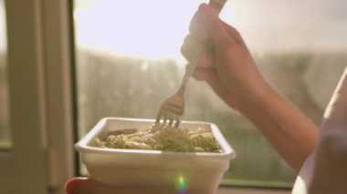 A image of someone eating instant noodles at home by the window in the kitchen, taken in a close-up shot. The fork is in the frame, along with the hands holding it, highlighting the act of consumption