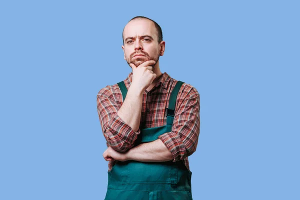 Meditative, handsome employee in overalls looking at camera. Serious, bearded man in studio shot. young workman seems lost in thought as he gazes off Dressed in overalls, , Doubt concept