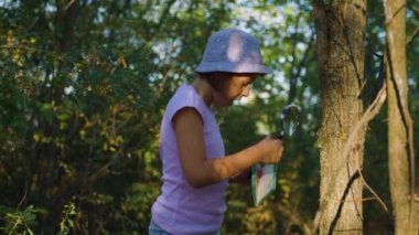 shocked kid girl exploring nature outside with magnifying glasses and makes notes in the notebook. in green forest. little girl looks through magnifying glass. children dreams of an adventure.