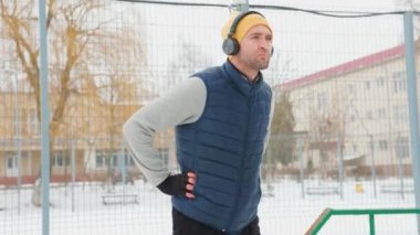 A young, athletic man exercises outside in the wintertime, wearing headphones and performing gymnastic movements. He embodies the epitome of a healthy lifestyle.