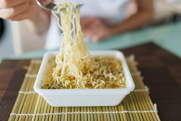 A shot of someone dining at home, with a fork in hand and a plate of instant noodles in front of them. with the focus on the fork in their hand.