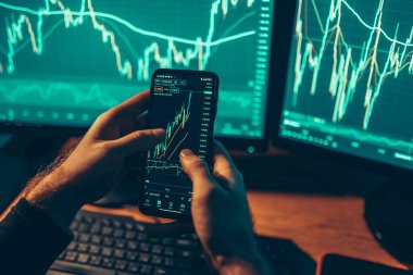 A close-up shot of his hands holding a smartphone and calculator, combined with the glowing computer screens, creates a mesmerizing scene of commerce and investment.