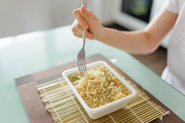 A shot of someone enjoying a quick and unhealthy meal of instant noodles, with the focus on the fork in their hand. with a fork in hand and a plate of fast food in front of them.