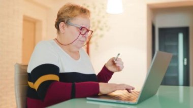 An overweight woman, wearing her eyeglasses, is shopping online from the comfort of her home. She is using her laptop and credit card to make purchases through various e-commerce sites,