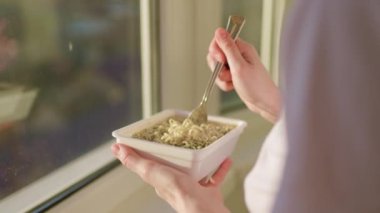 in a fast food meal of instant noodles at home by the window in the kitchen. The close-up shot highlights the use of a fork and the act of eating, emphasizing the consumption of the unhealthy meal.