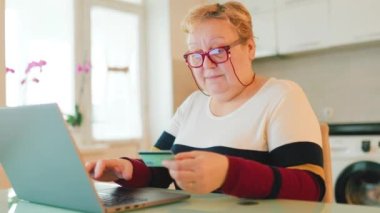 Şişman, yaşlı bir kadın yüzünde büyük bir gülümsemeyle elinde bir dizüstü bilgisayar ve kredi kartı ile online alışveriş yapmaktan memnun olduğunu gösteren bir ifade ile görülüyor..