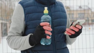 close up view of two gloved male hands with water bottle and smartphone in winter time