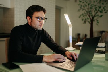 Online Job Young Male Professional Working Late with Laptop at Home. Serious student guy typing on his laptop and working late into the night at home.