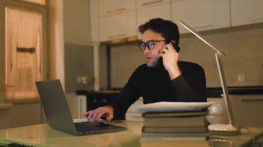 A freelance worker talking on the smartphone burning the midnight oil, typing away on his laptop as the night sky outside his window grows darker.