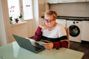A senior woman, looking overweight, is sitting at home and using her laptop while holding her credit card, making an online purchase through e-commerce.