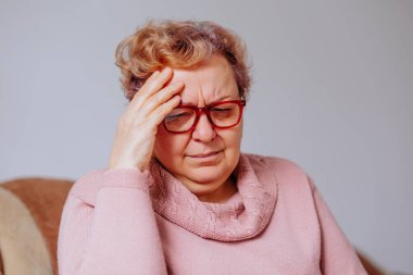 Exhausted senior woman with hands on head, grimacing in pain, experiencing a severe headache in the comfort of her own home