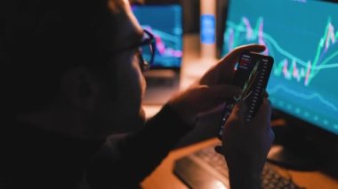 With a close-up shot of his hands holding a smartphone and calculator, the man works tirelessly through the night, analyzing charts and graphs displayed on the computer screen.