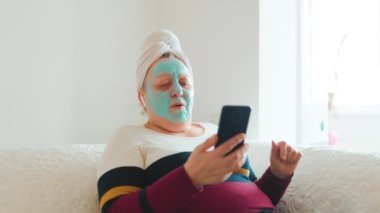 Dinlenme için müzik ve cilt bakımı. Oturma odasında oturan olgun bir kadın, yüzünde maske ve akıllı telefonunda çalan müzik eşliğinde rahatlatıcı bir cilt bakımına kendini kaptırıyor..
