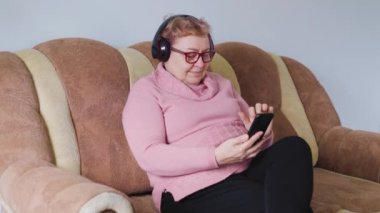 An elderly woman sitting comfortably on her sofa, listening to her favorite tunes with her headphones, looking happy and relaxed at home. Smiling Senior Lady Relaxing on Sofa with Headphones On