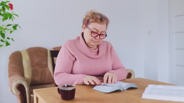 一位中年妇女坐在客厅的沙发上 脸上戴着眼镜 手里拿着一本小说 她全神贯注地看书 超重的身体舒舒服服地靠在垫子上 — 图库视频影像