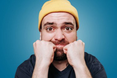 Şiddet dolu sakallı genç bir erkeğin portresi. Yüzündeki öfke doğrudan kameraya bakıyor. Mavi arkaplanda stüdyo çekimi,