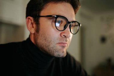 A young man in eyeglasses types on his laptop at home, with a gentle glow from the lamp lighting his face in the nighttime.