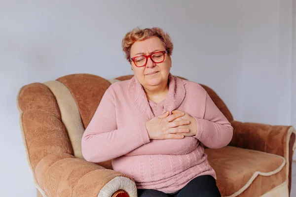 Overweight and Overwhelmed upset Woman Suffers Heart Attack on Sofa, an older woman is receiving treatment for acute heart pain at home. She is sitting on her sofa with a concerned expression