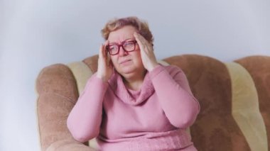 Exhausted senior woman with hands on head, grimacing in pain, experiencing a severe headache in the comfort of her own home