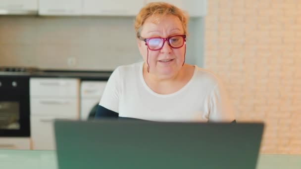 在家学习一个女人在家里 带着她的笔记本电脑和笔记本在网上学习 她是一个年龄较大的人 戴着眼镜 表明她积极参加了这个课程 — 图库视频影像