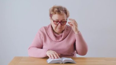 Tired and Sad Senior Woman at Home Struggling with Fatigue While Reading a Book. An old woman sits at home, reading a book with tired eyes and a look of fatigue on her face.