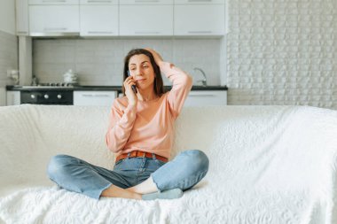 Neşeli genç bir kadın mutfak masasında oturuyor, akıllı telefonuyla ailesiyle konuşurken gülümsüyor. Otantik ve rahat atmosfer fotoğrafta yer alıyor.,