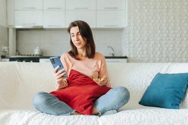 The young female sits cross-legged on the couch in her living room, deeply engrossed in surfing the net on her smartphone, with a stack of books on the side table beside her.