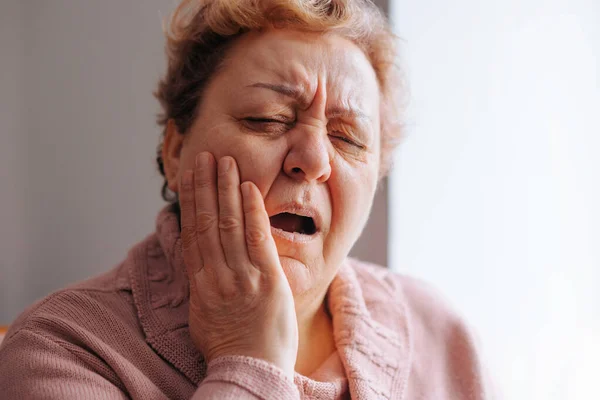 Aching Teeth Senior Woman in Pain at Home. A senior woman experiences a toothache while sitting at home, with a sad expression on her face.